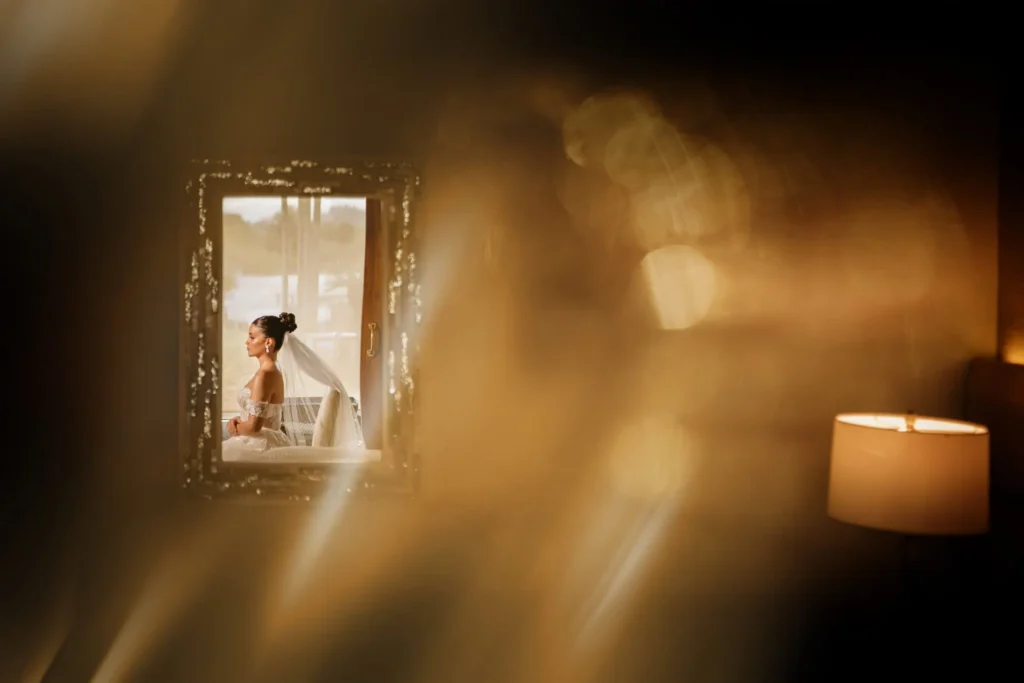 A bride in a white gown sits by a window in a softly lit room, gazing outward with a serene expression, warm bokeh lights adding romance in Medellín — Photographer John Palacio