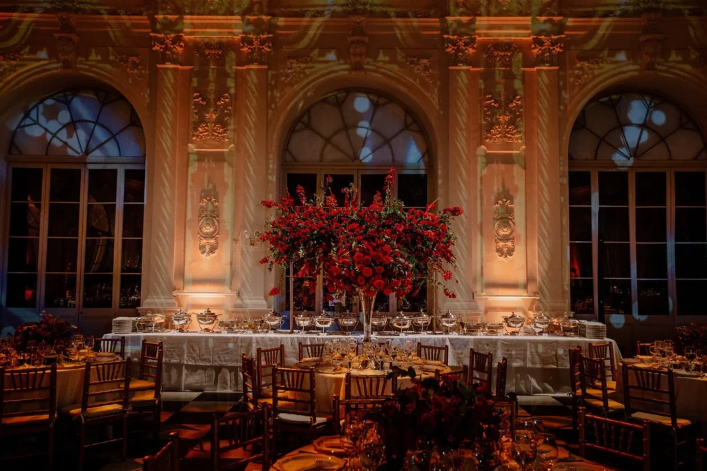 Luxurious wedding reception in Cartagena featuring tall red rose centerpieces on long tables, ornate arches, and warm lighting creating a romantic atmosphere in a grand hall — John Palacio