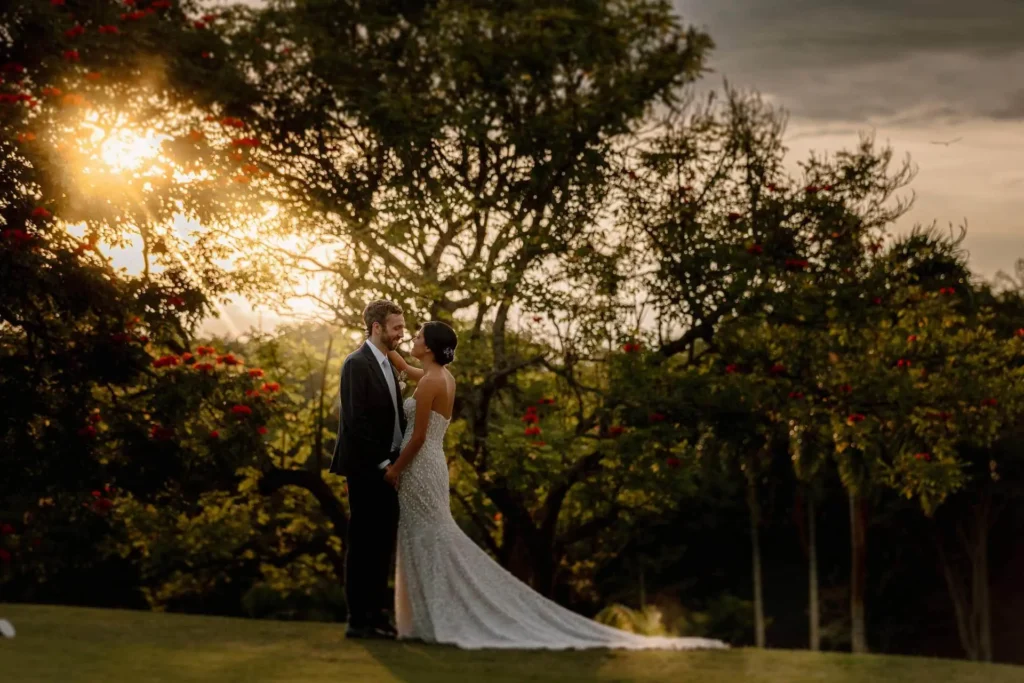 A bride in a flowing white gown and groom in a dark suit share an intimate embrace at sunset in a lush Medellín garden, with golden light filtering through red-flowered trees creating a warm, romantic mood — photographer John Palacio