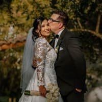 A joyful bride in lace gown and veil kisses her groom in tuxedo outdoors among lush trees in Cartagena, capturing tender wedding love and celebration — John Palacio