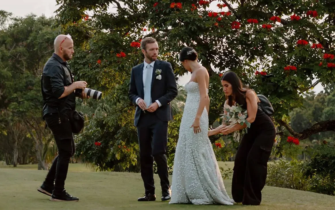 Bride in flowing white gown and groom in suit stand with photographer and assistant in lush tropical garden in Medellín, Colombia, amid red flowers and green foliage, evoking romantic wedding joy — John Palacio