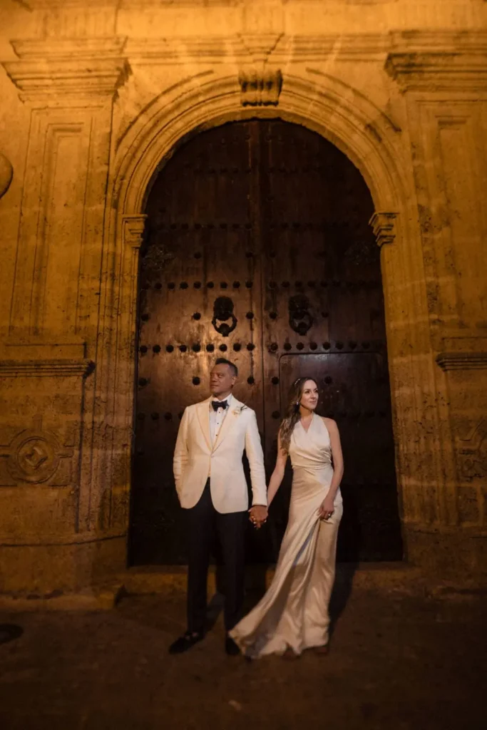 Una novia en vestido blanco fluido y novio en esmoquin blanco se toman de la mano ante una gran puerta de madera arqueada en el distrito histórico de Cartagena por la noche, evocando romance y elegancia — John Palacio