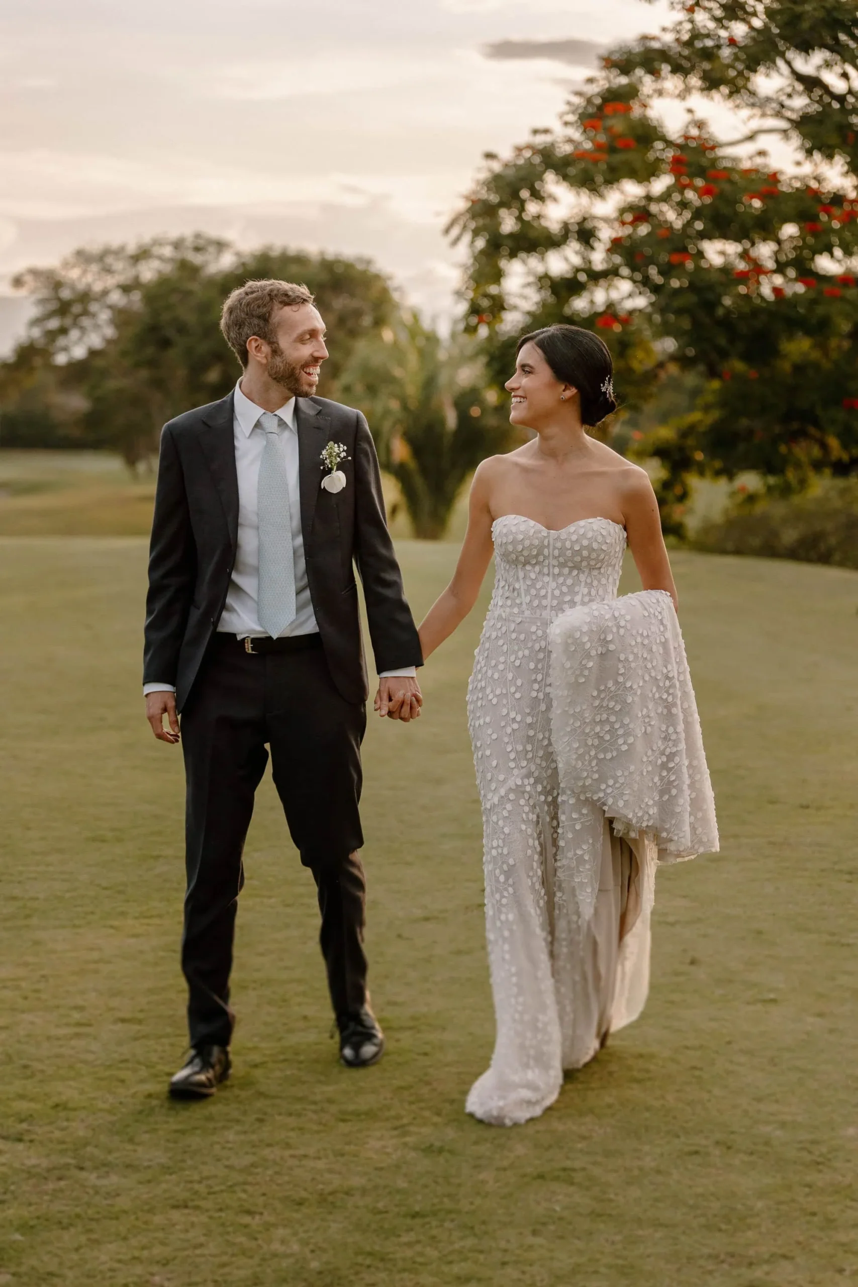 A joyful bride in a sparkling white gown and groom in a black suit hold hands while walking on a lush golf course at sunset in Medellín, capturing romantic wedding vibes — John Palacio