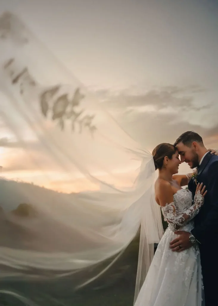 Romantic bride and groom embrace outdoors at sunset in Medellín, with her lace gown and flowing veil against hilly backdrop, capturing tender wedding moment by Medellín wedding photographer — John Palacio