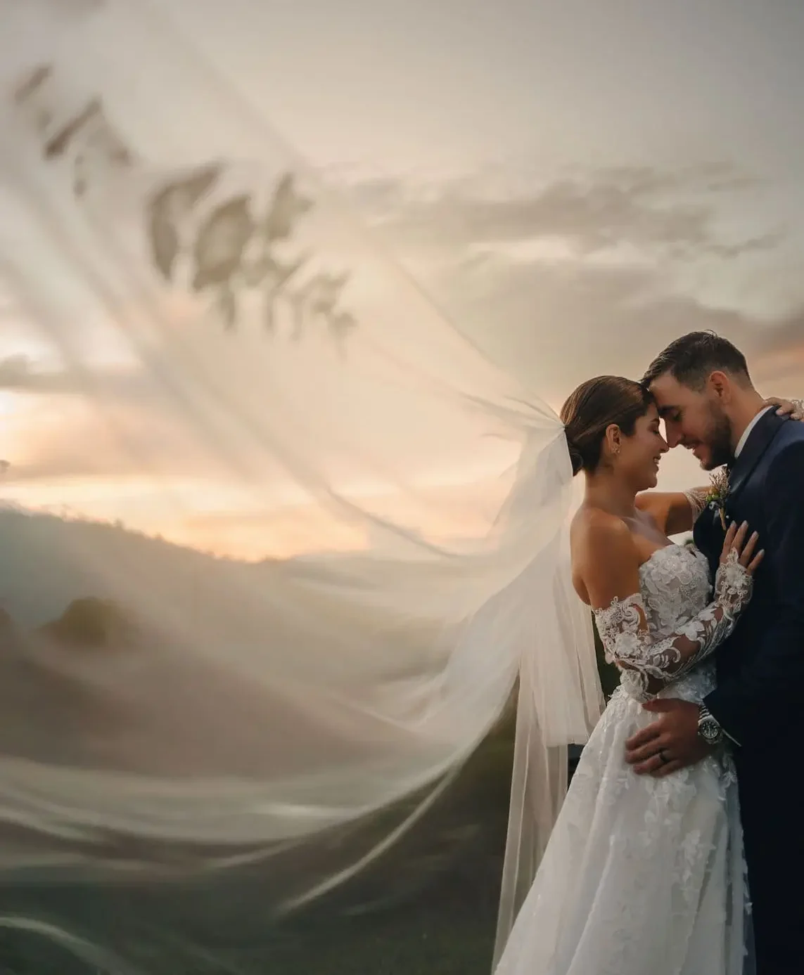 Novia y novio románticos se abrazan al aire libre al atardecer en Medellín, con su vestido de encaje y velo ondeante contra fondo montañoso, capturando tierno momento de boda por fotógrafo de bodas en Medellín — John Palacio